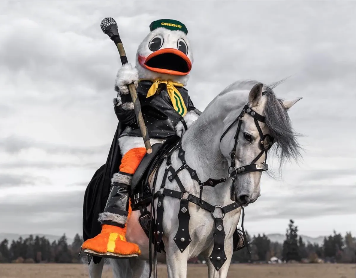 The Oregon Duck in a studded black jacket and cape, holding a staff and riding a gray horse in black leather tack under a cloudy sky — a still from Game of Ducks.